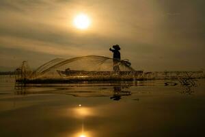 silhouette di pescatore a Alba, in piedi a bordo un' canottaggio barca e getto un' netto per catturare pesce per cibo foto