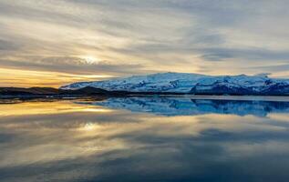 lago e nevoso montagne a tramonto nel Islanda con maestoso nordico paesaggio, congelamento freddo acqua. scandinavo ciglio della strada scenario con colline durante d'oro ora, Paese delle meraviglie panoramico percorso. foto