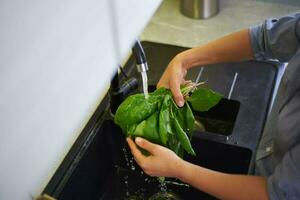 alto Visualizza di donna di mani lavaggio fresco biologico spinaci le foglie sotto fluente acqua nel il cucina lavello. foto