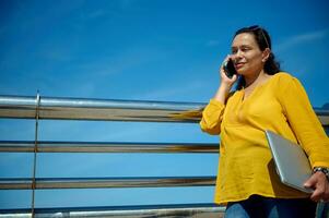 fiducioso donna nel giallo casuale camicia, in piedi di metallo recinto con un' il computer portatile nel sua mano, parlando su inteligente mobile Telefono foto