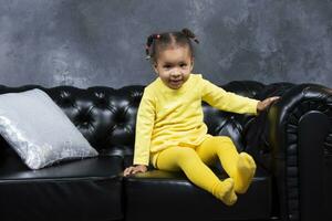 poco africano americano ragazza si siede su un' nero pelle divano. foto
