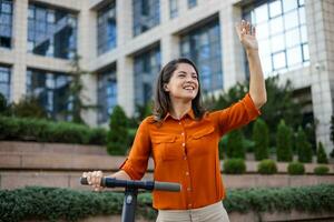 giovane bellissimo donna sorrisi e cavalcate un elettrico scooter per opera lungo ufficio edifici. ecologico mezzi di trasporto concetto. foto