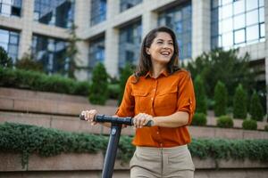 giovane bellissimo donna sorrisi e cavalcate un elettrico scooter per opera lungo ufficio edifici. ecologico mezzi di trasporto concetto. foto