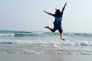 felicità di asiatico donna è salto su cielo con mare e onda su il sabbia spiaggia nel naturale tramonto. la libertà solitario vita su vacanza foto