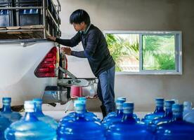 lavoratori sollevamento potabile acqua chiaro e pulito nel blu plastica gallone in il indietro di un' trasporto camion purificato potabile acqua dentro il produzione linea per preparare per vendita.piccolo attività commerciale foto