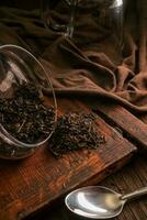 foglia delizioso secco tè su un' di legno Marrone tavolo con un' cucchiaio, stoffa e cristalleria su un' Marrone buio sfondo foto