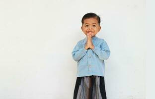 un' carino sorridente poco ragazzo con blu camicia e sarong saluto qualcuno foto