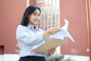 sorridente asiatico donna d'affari manager lettura rapporto carta mentre a piedi nel all'aperto, contento commessa supervisore verivy i saldi dati mentre a piedi al di sopra di città edificio foto