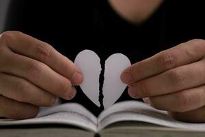 femmina mano Tenere un' mezzo strappato bianca carta cuore al di sopra di un Aperto libro su il tavolo. bianca carta cuore strappato nel metà nel mano foto