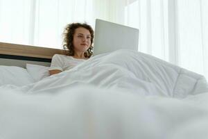 giovane bellissimo contento donna rilassante e Lavorando con il computer portatile mentre dire bugie nel letto nel il mattina dopo veglia su. foto