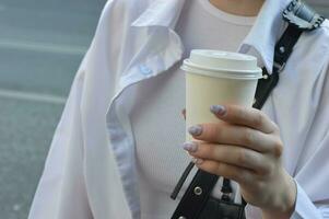 ragazza nel un' bianca camicia con un' bicchiere di caffè nel sua mano. un' donna di mano detiene un' monouso carta tazza. avvicinamento. foto