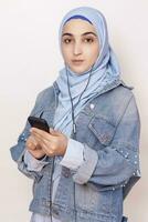 moderno musulmano ragazza ascolta per musica, podcast, e audiolibri su smartphone. contento musulmano donna nel hijab ascoltando canzoni con cuffie foto