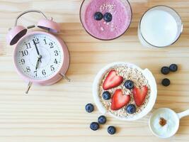 piatto posare di allarme orologio, prima colazione con avena o muesli nel bianca ciotola, fresco mirtilli, fragole, un' bicchiere di latte, mirtillo frullato e Yogurt su di legno tavolo. salutare prima colazione concetto. foto