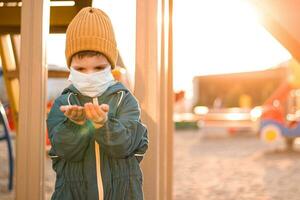 un' ragazzo nel un' protettivo maschera giochi nel il terreno di gioco e fuoriuscite sabbia a partire dal il suo mani durante il pandemia di coronavirus e covid - 19 foto