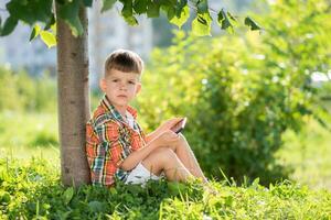 un' bellissimo bambino si siede su il erba e sembra a il Telefono nel il estate a tramonto. carino ragazzo con mobile nel mano foto