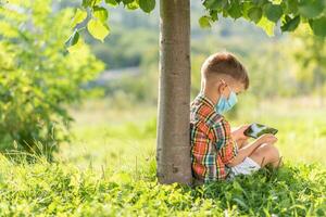 un' ragazzo nel un' medico maschera si siede su il erba e sembra nel il Telefono cartoni animati nel il estate a tramonto. bambino con un' mobile Telefono nel il suo mani. prevenzione contro coronavirus covid-19 durante un' pandemia foto