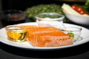 vicino su per salmone trota bistecca, crudo filetto di mare pesce su bianca piatto con sale, olio, e wasabi crema nel studio leggero su buio sfondo. foto