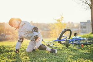 un' piccolo bambino nel un' casco e protezione abbattere a partire dal un' bicicletta su il erba e era non ferito foto
