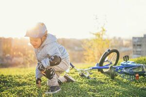 un' piccolo bambino nel un' casco e protezione abbattere a partire dal un' bicicletta su il erba e era non ferito foto