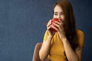 Sorridi giovane asiatico donna Tenere un' rosso tazza e godendo potabile caldo caffè nel il mattina, concetto di rilassamento nel tempo libero e merce pasto prima colazione a casa. foto