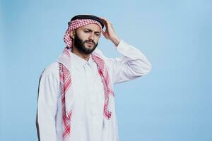 esausto musulmano uomo vestito nel tradizionale Abiti Tenere testa nel dolore mentre sofferenza a partire dal emicrania. stanco arabo persona indossare islamico thobe e foulard avendo mal di testa foto