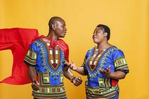 africano americano donna gesticolando e guardare a uomo vestito nel superuomo mantello. sorridente fidanzato indossare rosso supereroe mantello avendo comunicazione con fidanzata nel studio foto