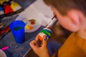 un' bambino decora un Pasqua uovo nel il colori di il arcobaleno. un' bambino detiene un uovo e vernici esso con un' spazzola. preparazione per il celebrazione di Pasqua. foto