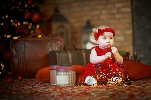 poco ragazza nel rosso vestito contro sfondo di Natale albero detiene Natale ghirlanda nel sua mani. bambino 6 mese vecchio celebra Natale. foto