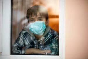 bambino nel protettivo medico maschera sembra su di il finestra in strada. restare casa. solitudine durante coronavirus epidemico. scuola quarantena. foto