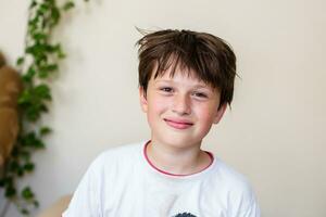 bellissimo adolescente ragazzo a casa contro leggero parete con tessitura impianti. ragazzo nel bianca maglietta e con spettinato capelli sorrisi. divertimento le persone. foto