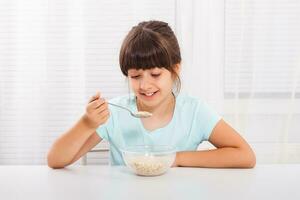 carino poco ragazza è seduta a il tavolo e mangiare cereali per prima colazione. foto