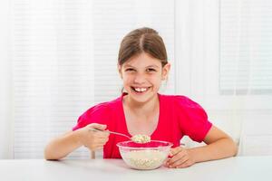 carino poco ragazza è seduta a il tavolo e mangiare cereali per prima colazione. foto