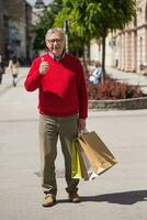 anziano uomo mostrando pollice su mentre gode nel shopping a il città foto