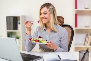 bellissimo donna d'affari gode mangiare insalata mentre Lavorando nel sua ufficio foto