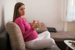 triste incinta donna potabile pillola mentre seduta su divano a sua casa. foto