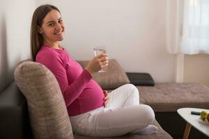 bellissimo incinta donna gode potabile acqua mentre riposo su divano a sua casa. foto