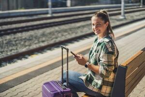 contento donna utilizzando mobile Telefono mentre seduta su un' panchina a il treno stazione foto