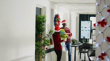 dipendenti nel Natale decorato ufficio godendo tazza di caffè di il acqua più fresco, ridendo insieme. contento bipoc personale membri indossare Santa Claus cappelli assunzione rompere nel multirazziale spazio di lavoro foto