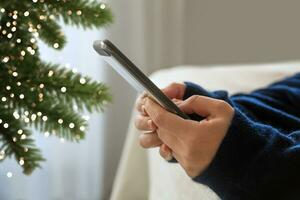 ragazza mani con un' smartphone menzogna su il letto e un' Natale albero con un' ghirlanda.bokeh. selettivo messa a fuoco. alto qualità foto