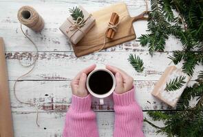 mani nel rosa maglione hold bianca tazza di caffè su un vecchio di legno opera tavolo con fatto a mano i regali e rami di Natale albero.piatto lay.top Visualizza foto