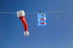 avvicinamento poco Santa Claus cappello e un' credito carta con un' immagine sospeso su un' corda contro il blu cielo. avvicinamento, allegro Natale celebrazione, pubblicità, bigboard foto