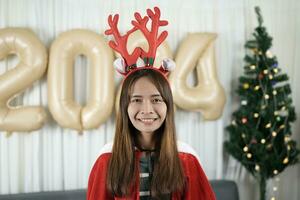 asiatico donna vestito come un' baracca sorridente felicemente in attesa per celebrare Natale 2024. decorare e decorare il camera con un' Natale albero e 2024 palloncini. foto