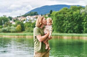 ritratto di contento giovane padre con adorabile bambino piccolo ragazza giocando al di fuori Il prossimo per lago o fiume foto