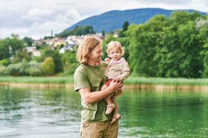 ritratto di contento giovane padre con adorabile bambino piccolo ragazza giocando al di fuori Il prossimo per lago o fiume foto