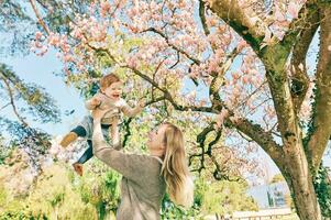 all'aperto ritratto di contento giovane madre con adorabile bambino ragazza sotto fioritura primavera albero, avendo bene tempo nel soleggiato parco foto