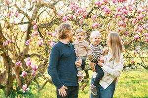 all'aperto ritratto di contento giovane famiglia giocando nel primavera parco sotto fioritura magnolia albero, bello coppia con Due poco bambini avendo divertimento nel soleggiato giardino foto