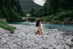 giovane donna è seduta su il pietra nel il mezzo di un' torrente. paesaggio con azzurro pittoresco sorprendente montagna lago. su il riva si siede un' misterioso brunetta ragazza con lungo capelli. Baviera. Austria foto