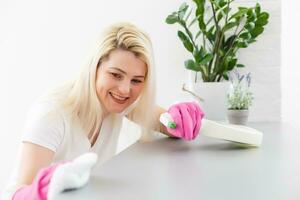 donna nel protettivo guanti è sorridente e asciugandosi polvere utilizzando un' spray e un' spolverino mentre pulizia sua Casa, avvicinamento foto