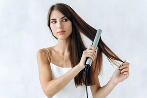 primo piano ritratto di una giovane donna sorridente in abiti domestici utilizzando una piastra per capelli mentre si prepara a camminare. concetto di stile di vita domestico foto