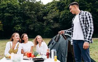 uomo in piedi accanto a una tenda che tiene una birra e guarda le donne all'aperto nel campeggio foto